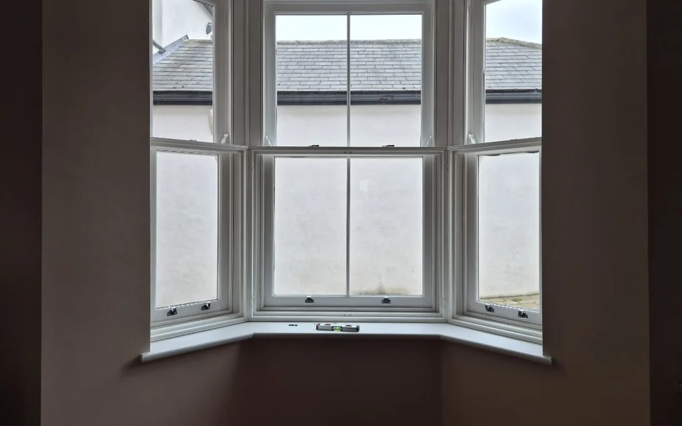 Three-part white sash bay window in Aberford before shutters, lower sashes with privacy glazing and a spirit level on the sill.