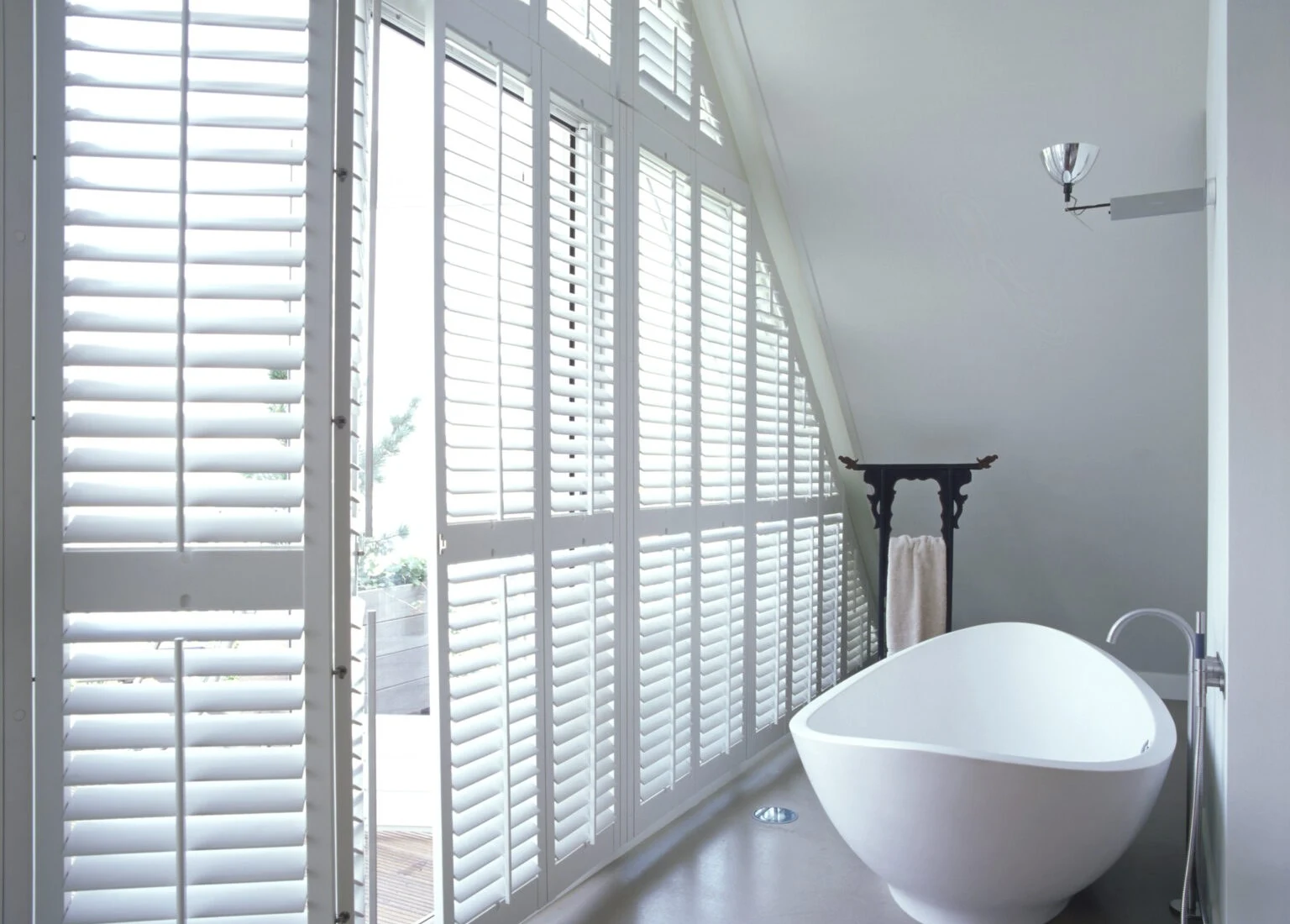 Full-height white Java shutters on a sloping bathroom window wall beside a freestanding bath.