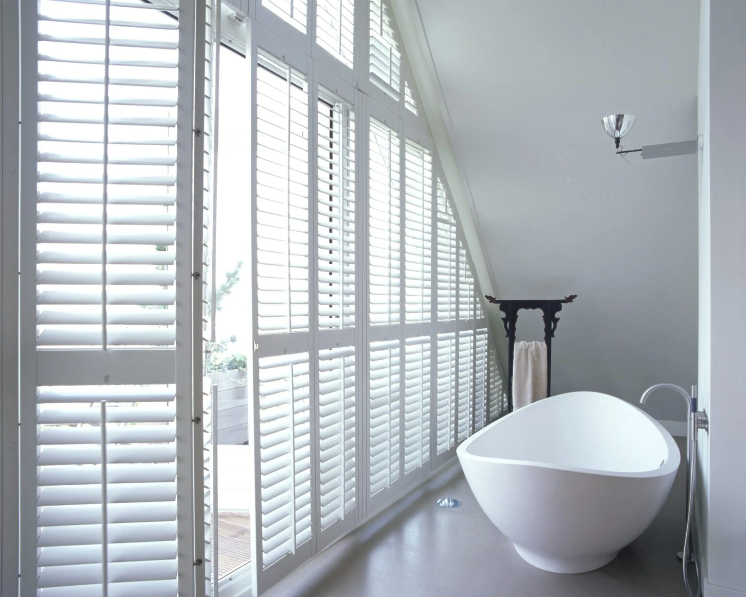 Full-height white Java shutters on a sloping bathroom wall above a freestanding bath.