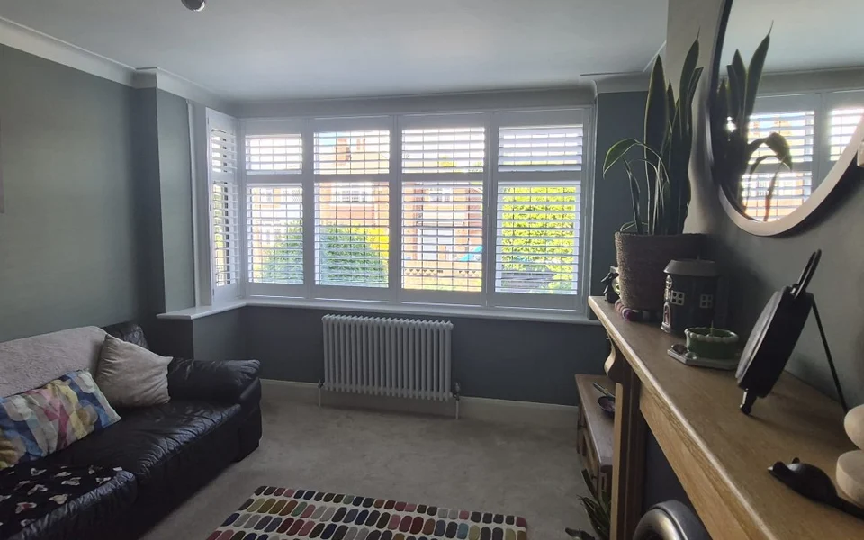 Same Bolton lounge bay after installation of white full-height plantation shutters with louvres tilted open across five panels.