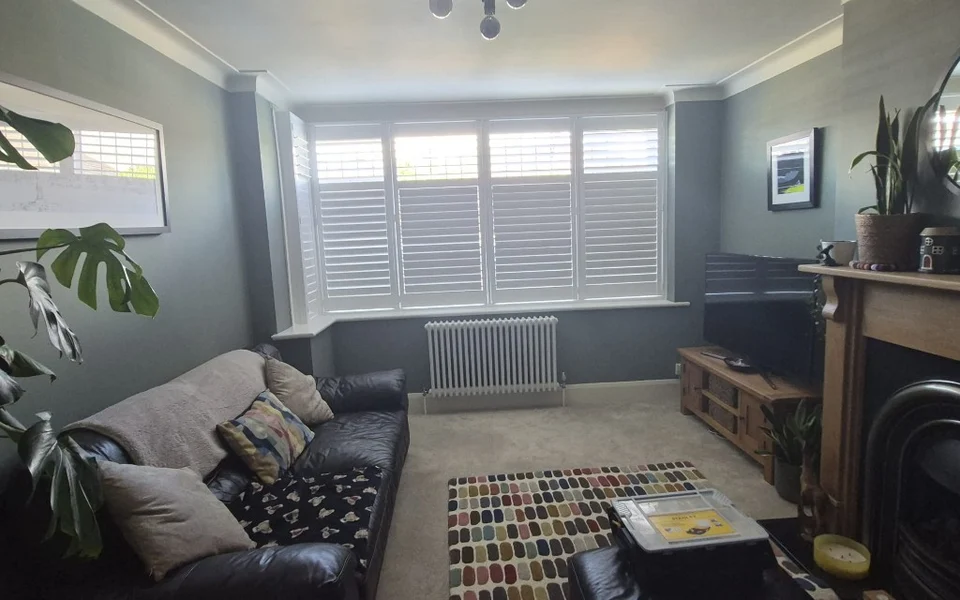 Wide interior shot of Bolton lounge with bay window shutters, leather sofa, fireplace, and patterned rug.