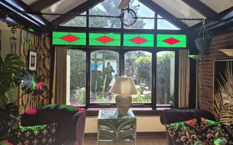Castleford conservatory before shutters, showing four arched windows under green and red stained-glass panels with purple armchairs and a Monstera plant.