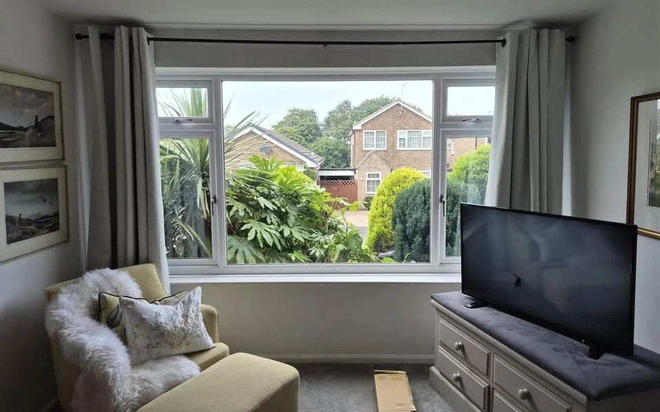 Catterick lounge before shutters, showing a wide three-pane window with curtains pulled back, garden view beyond, and a cardboard box on the carpet.