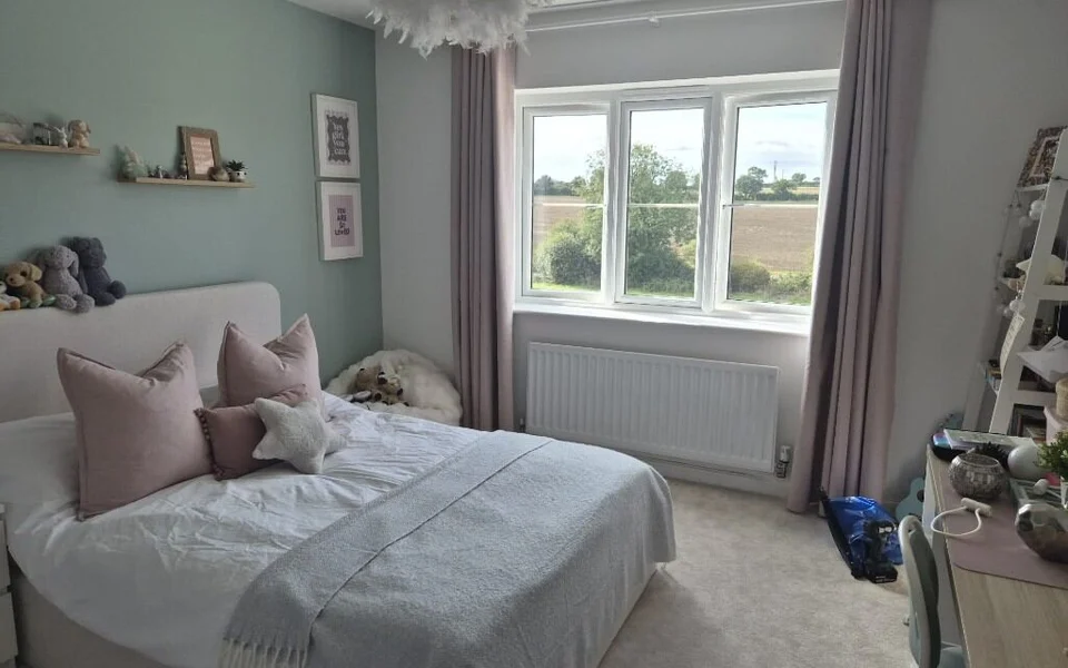 Garforth bedroom before shutters, a three-pane window with mauve curtains, a pale green feature wall and a double bed with pink cushions.