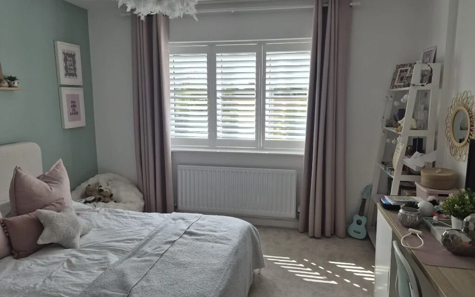 Same Garforth bedroom after white plantation shutters were fitted across the three-pane window, curtains kept either side and light patterns on the carpet.