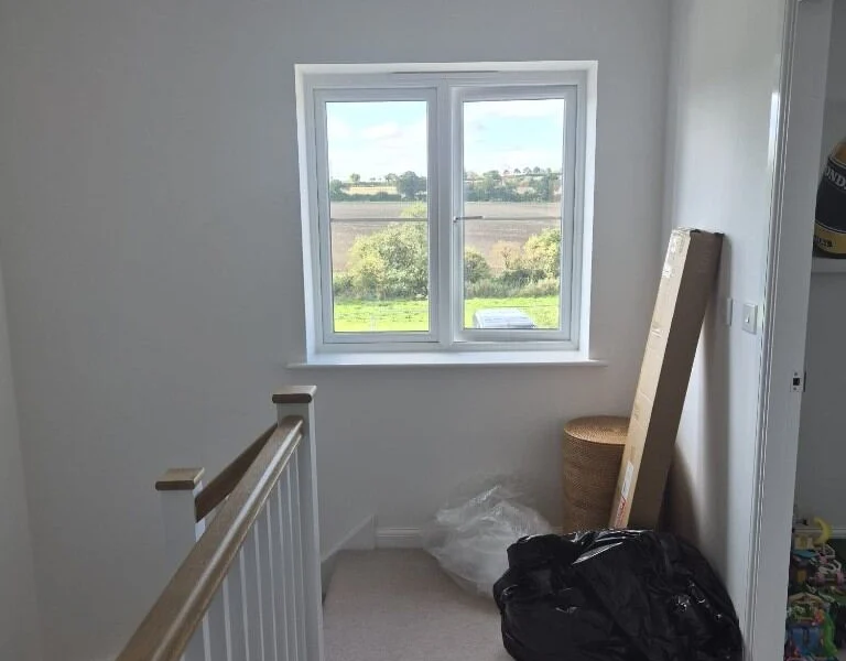 Garforth stair landing before shutters, a bare two-pane window overlooking open fields above an oak and white banister.
