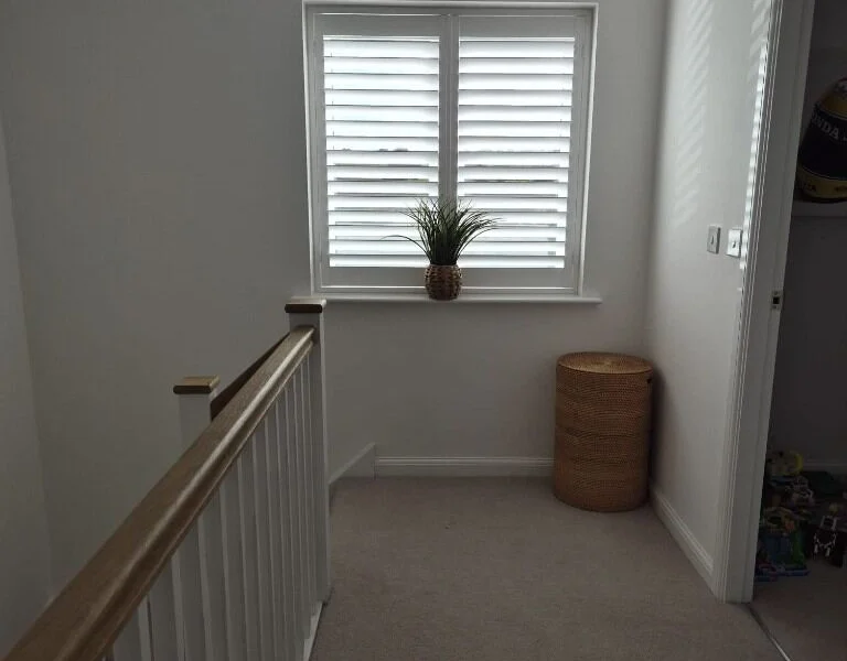 Same Garforth landing after two white plantation shutter panels were fitted in the window, with a small plant on the sill.