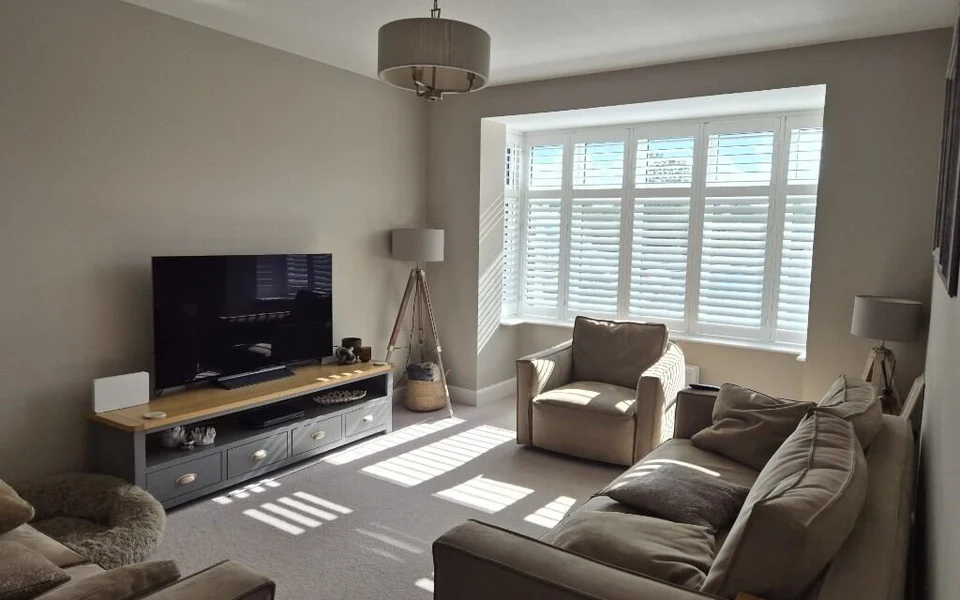 Garforth lounge after white plantation shutters were fitted across a bay window, sunlight striping the carpet between the sofas and TV unit.