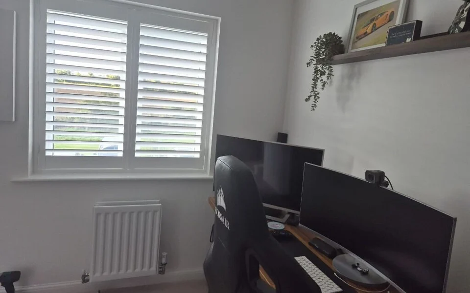 Garforth home office after white plantation shutters were fitted to a two-pane window beside a desk with monitors and a gaming chair.