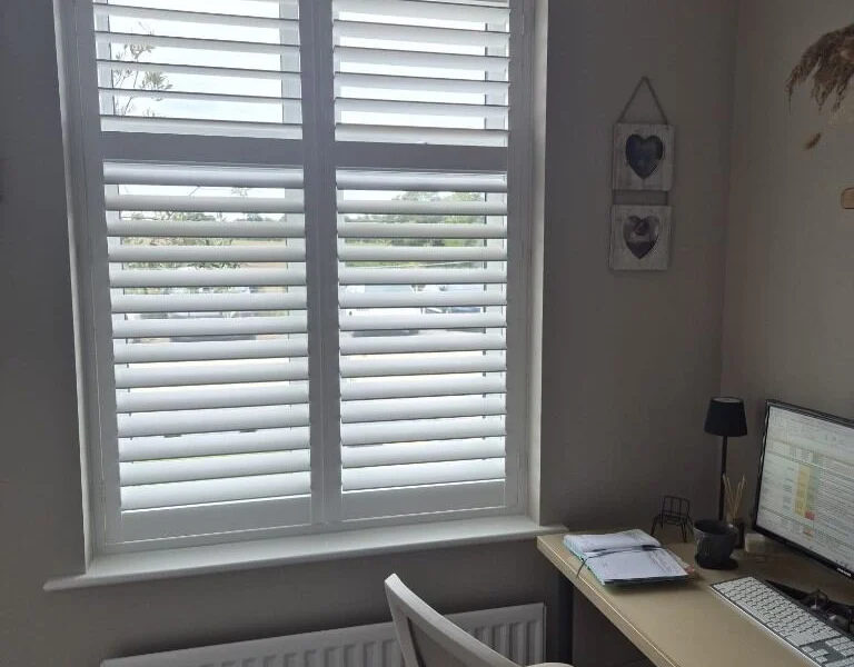 Second Garforth home office after white plantation shutters were fitted to a tall window with a transom, next to a desk and white office chair.