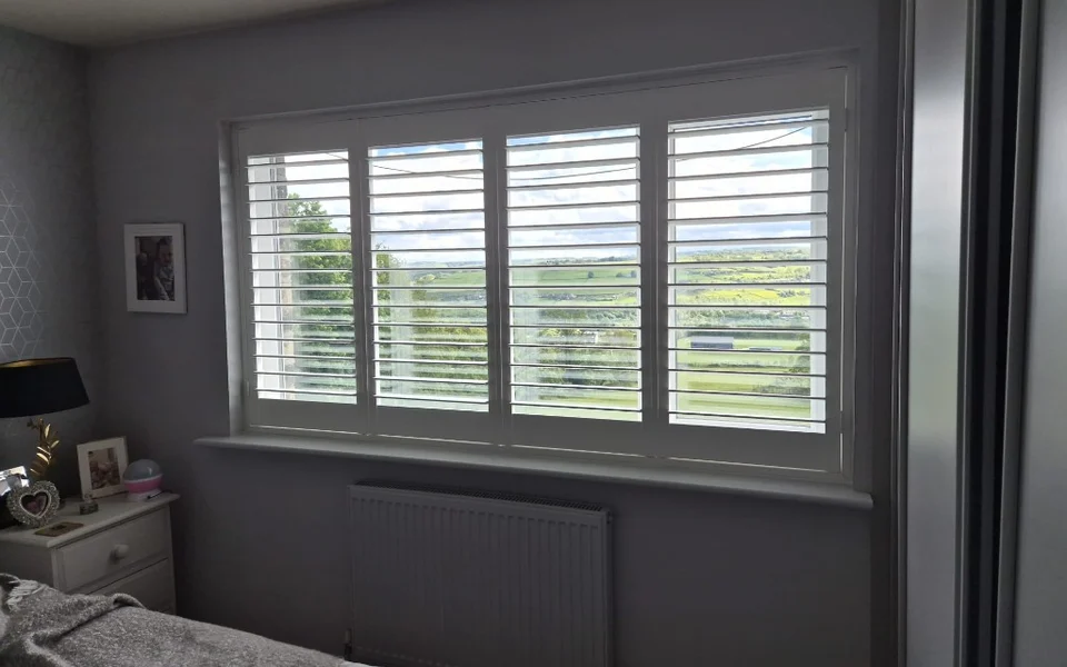 Halifax bedroom with white full-height plantation shutters on a wide window, louvres open to the view.