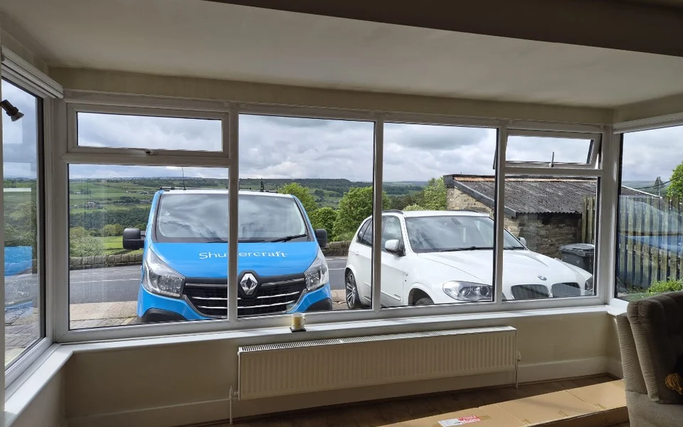 Halifax lounge bay window before shutters, white uPVC frames with countryside views and shutter packaging on the floor.