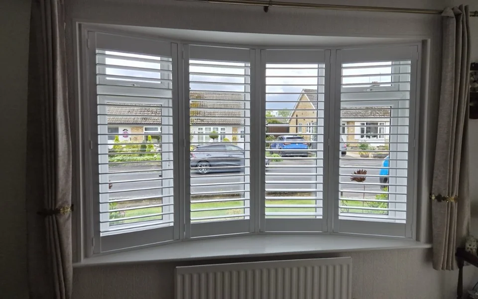 Harrogate lounge bay with white plantation shutters, louvres open, with curtains tied back.