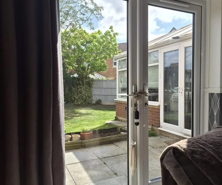 Harrogate lounge patio doors before shutters, white uPVC double doors open to a paved garden with a dark curtain to one side.