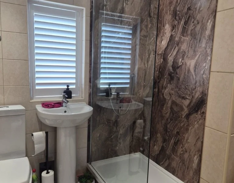 Huddersfield bathroom with beige tiles and a small window fitted with white plantation shutters beside a pedestal sink.