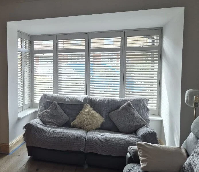 Huddersfield lounge after light grey plantation shutters were fitted across a deep bay, behind a grey sofa with cushions.