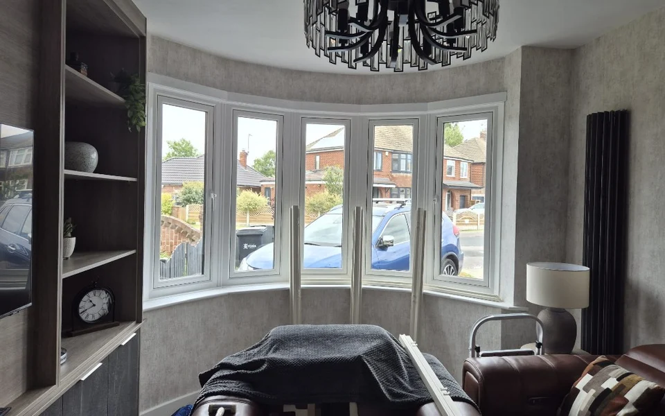 Huntington lounge bay before shutters, five-panel white bay window with shutter components leaning against the glass and a brown leather sofa.