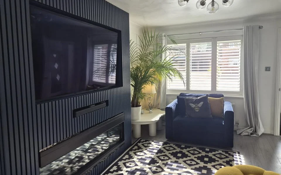 Leeds lounge after white full-height plantation shutters were fitted on the window, with grey curtains, a navy armchair, and geometric rug in daylight.