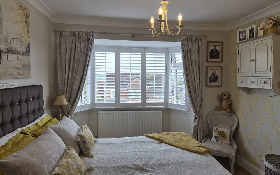 Pontefract bedroom bay with white plantation shutters, patterned curtains, grey upholstered headboard, and chandelier.