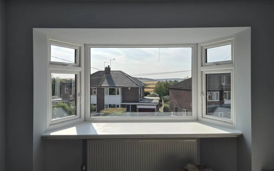 Pontefract display bay before shutters, three-section white uPVC bay with a deep sill and radiator below, looking out to brick houses and hills.