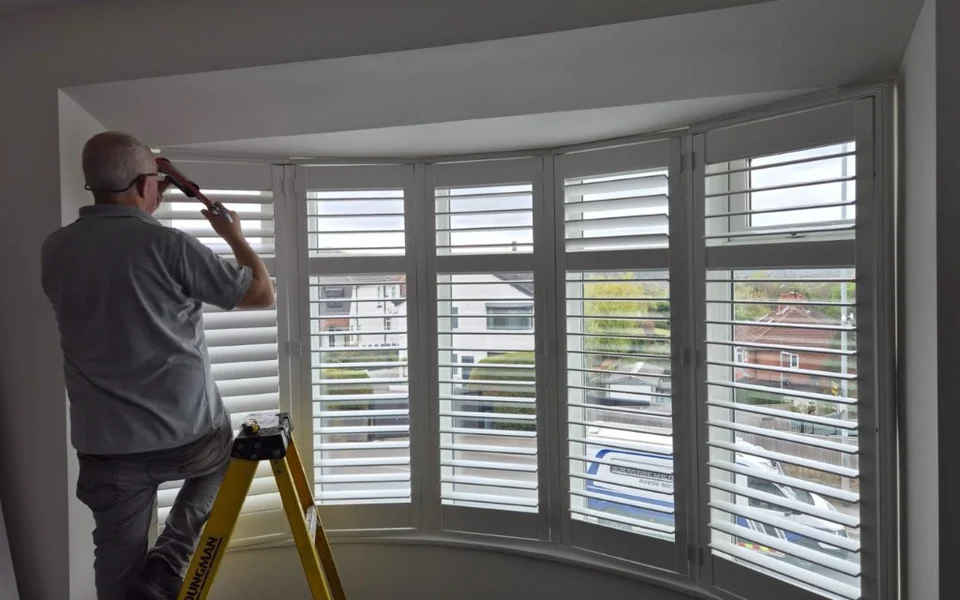 ShutterLuxe installer on a ladder sealing the frame of white plantation shutters on a large curved bay window in Rawdon.