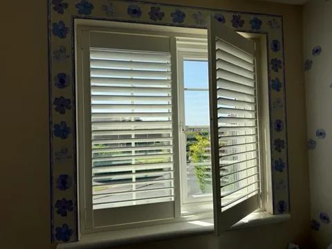 White plantation shutters with one panel hinged open to show the window behind