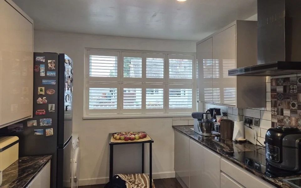 Wakefield galley kitchen after white tier-on-tier plantation shutters were fitted across a three-light window, between a fridge covered in magnets and light gloss units.