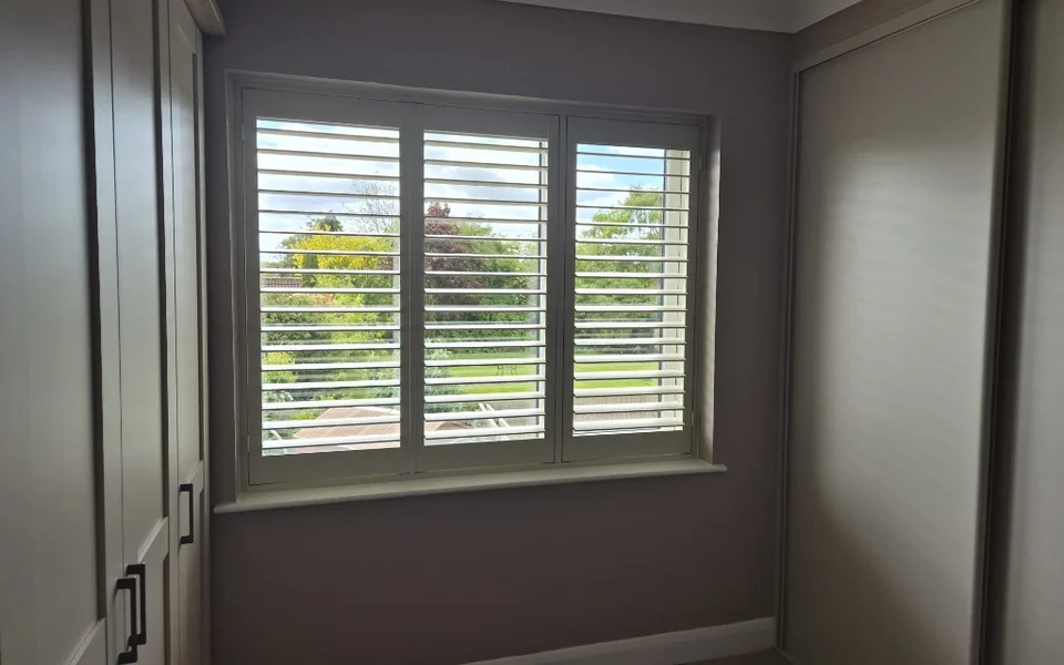 York bedroom with off-white full-height plantation shutters on a three-panel window, louvres open to a green garden view.